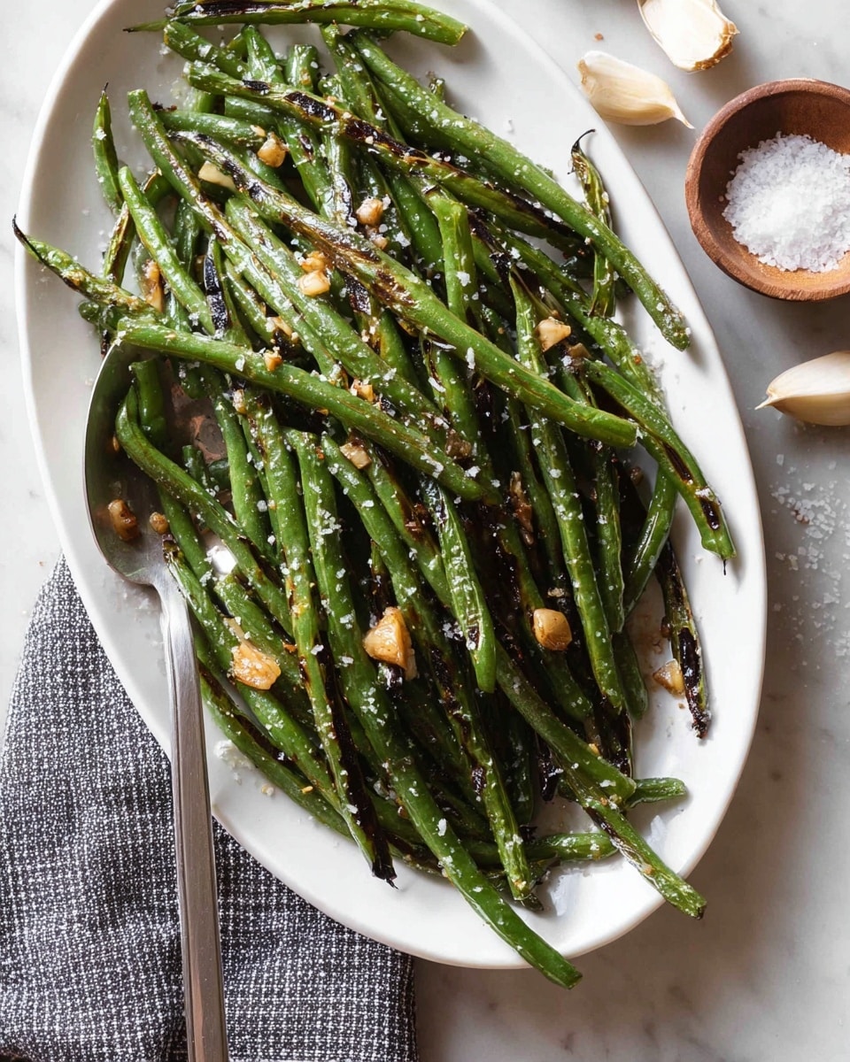 On a white oval plate, there is one layer of cooked green beans that are slightly charred with dark grill marks, giving a mix of bright green and deep brown colors. Small pieces of golden-brown garlic are scattered over the beans, along with coarse salt flakes for texture. A silver spoon rests on the left side of the plate. The plate is placed on a white marbled surface, with some coarse salt in a small wooden bowl and scattered garlic cloves nearby. A gray and white checkered cloth is partially visible at the bottom left corner. photo taken with an iphone --ar 4:5 --v 7