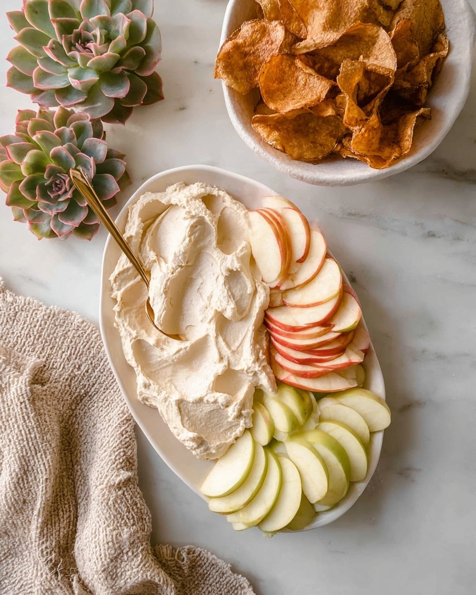 The image shows a white oval plate with two main layers. On the left side, there is a thick layer of creamy, off-white dip with a smooth and slightly fluffy texture. On the right side of the plate, there are two types of sliced apples: one with red skin and pale yellow inside, and another with pale greenish-yellow skin and inside, arranged in overlapping layers that cover about half the plate. A small gold spoon rests on the plate, partially immersed in the creamy dip. In the background, on a white marbled surface, there is a white bowl filled with large, brown, crispy chips, with a few scattered chips beside it. Surrounding the plate are some greenish-pink succulent plants and a light beige knitted cloth. Photo taken with an iphone --ar 4:5 --v 7