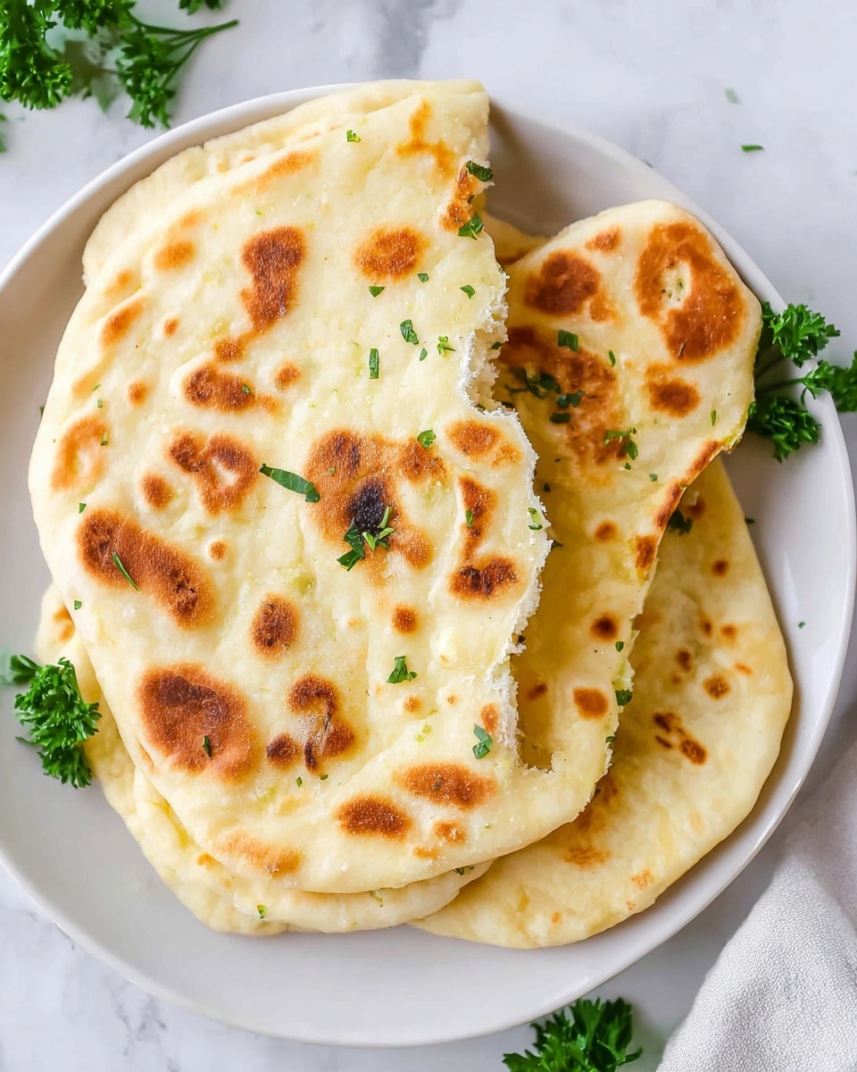A white plate holds four pieces of golden brown flatbread stacked irregularly, with the top piece torn to show a soft, fluffy inside. The flatbread has a warm, light yellow color with scattered darker brown spots from cooking, giving a textured look. Small bits of green herbs are lightly sprinkled over the top piece, adding a fresh touch. The plate is set on a white marbled surface with a few green parsley leaves around the edges. Photo taken with an iphone --ar 4:5 --v 7