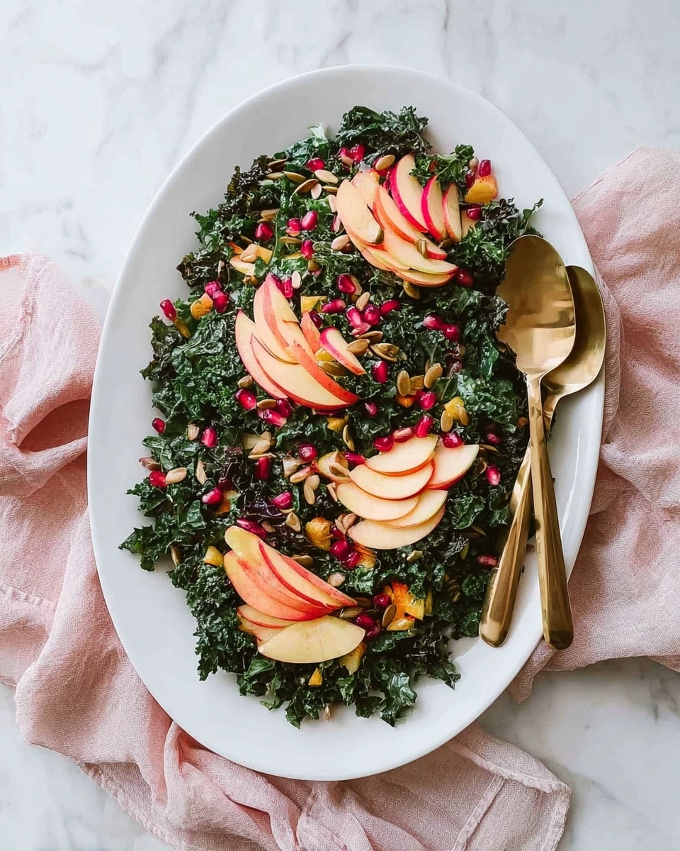 An oval white plate holds a fresh salad with a base layer of dark green chopped kale, covering the entire plate. Scattered evenly over the kale are small red pomegranate seeds and light brown pumpkin seeds, adding pops of color and texture. On top, four fan-shaped groups of thinly sliced apple pieces are arranged in a circle, their pale pink and yellow skin visible, adding a soft contrast to the dark greens. To the right side of the plate, two gold-colored serving spoons rest partly on the salad and partly on the edge. The plate is set on a white marbled surface with a soft pink cloth casually placed nearby. photo taken with an iphone --ar 4:5 --v 7