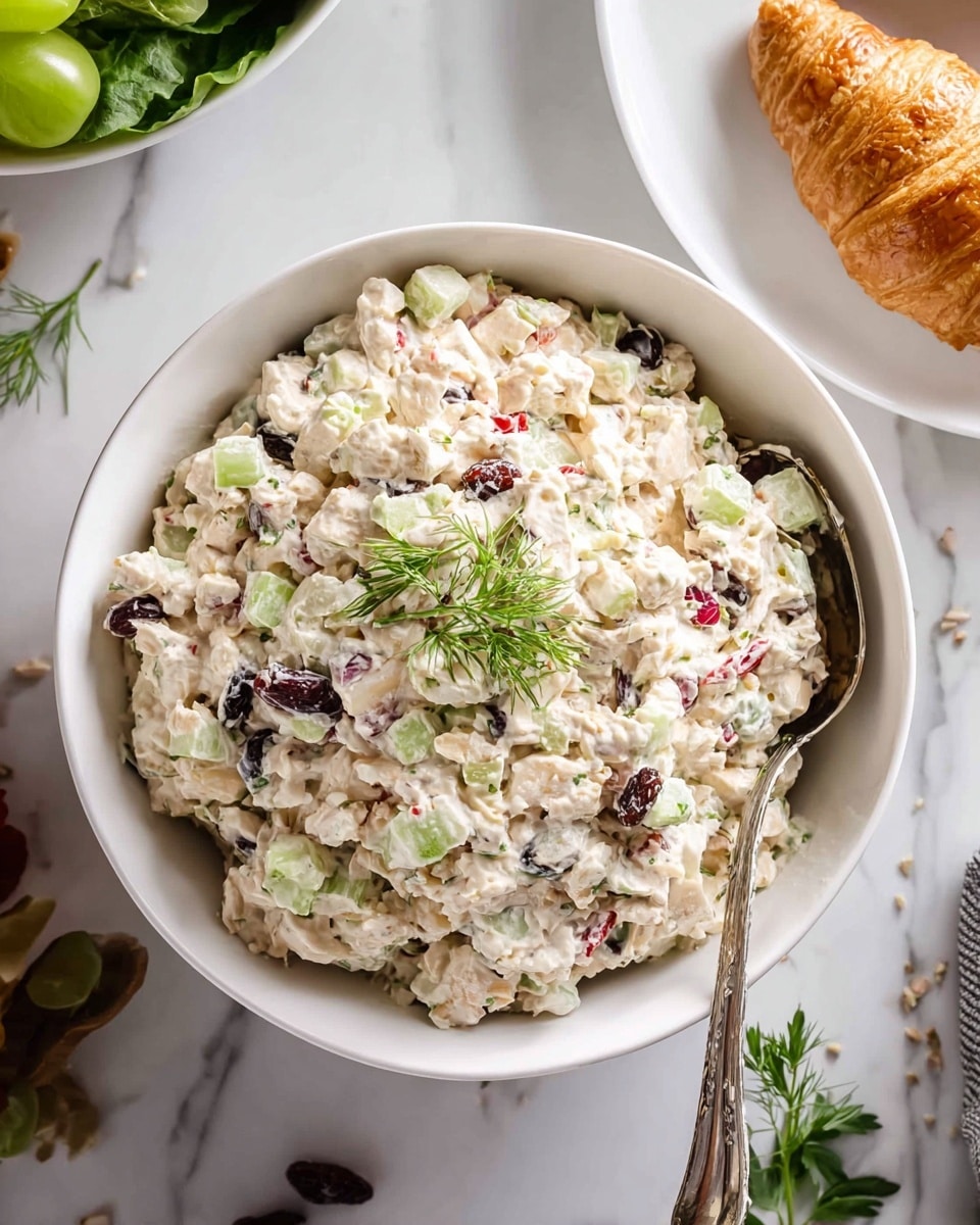 A white bowl is filled with a chunky salad that has about three layers of mixed ingredients. The base layer is creamy, pale off-white, mixed with small bits of light green celery, small red pieces that look like bell pepper, and small dark purple dried fruit. There are also small chunks of white, firm-looking apple or cheese, mixed with the creamy base and scattered evenly. On top, fresh green herb sprigs sit as decoration. A silver spoon rests on the right side inside the bowl. The bowl is placed on a white marbled surface with a croissant on a white plate and a bowl of fresh green leaves nearby. photo taken with an iphone --ar 4:5 --v 7