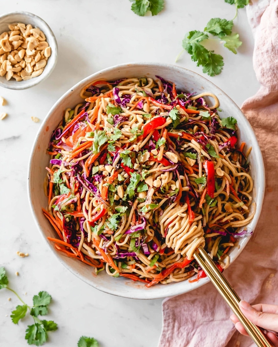 A large white bowl full of an Asian noodle salad sits on a white marbled surface. The dish has three main layers: the bottom is light brown thick noodles, mixed with thin shredded purple cabbage, bright orange carrot sticks, and thin sliced red bell peppers. On top, green cilantro leaves and light brown chopped peanuts are scattered, adding texture and color. A woman's hand holds gold chopsticks, pulling noodles from the right side of the bowl. Around the bowl are a small white bowl with peanuts, scattered cilantro leaves, and a light pink cloth. Photo taken with an iphone --ar 4:5 --v 7