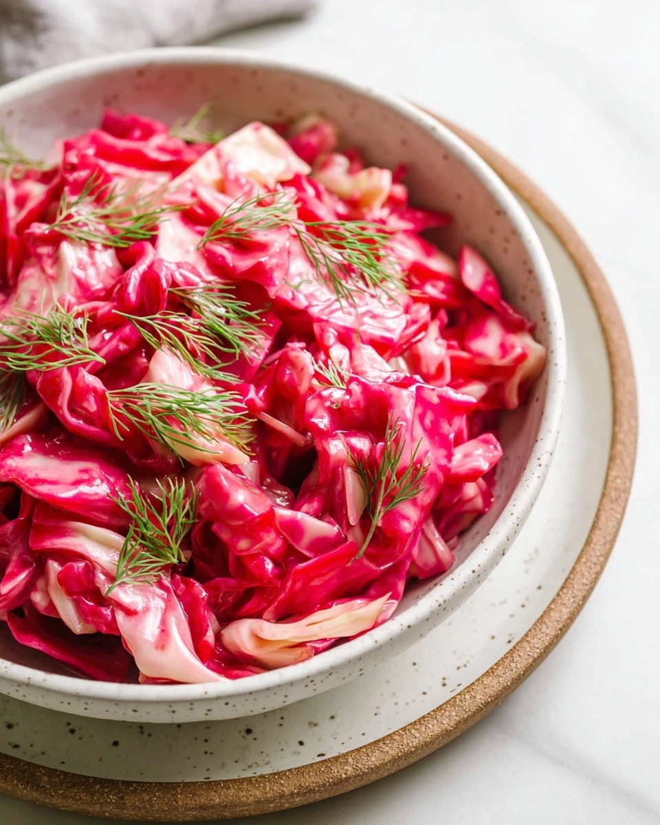 A close-up of a textured white bowl filled with a bright pink and light cream cabbage salad, where the cabbage pieces are roughly chopped and coated in a glossy, vibrant pink dressing. Light green dill sprigs are scattered on top of the salad, adding a fresh contrast. The bowl sits on a white plate with brown speckles and a light brown rim, all placed on a white marbled surface. Photo taken with an iphone --ar 4:5 --v 7