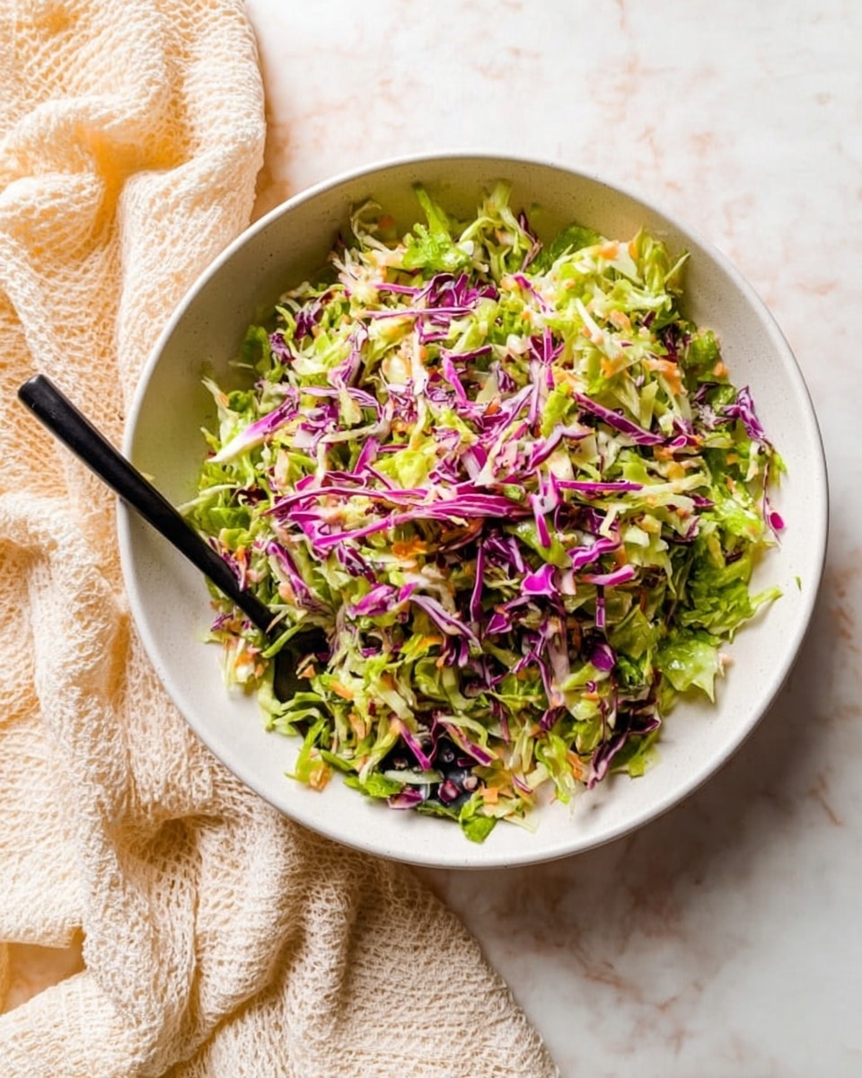 A dish of shredded salad with two visible layers, placed in a white bowl. The bottom layer is finely shredded bright green lettuce, mixed with some tiny bits of light orange and translucent white. On top is a sprinkle of thin purple cabbage strips adding color contrast. A black spoon rests near the edge of the bowl, partially buried in the salad. The bowl sits on a white marbled surface with a cream-colored textured cloth slightly under and around one side. The overall look is fresh and colorful with a natural light setting. photo taken with an iphone --ar 4:5 --v 7