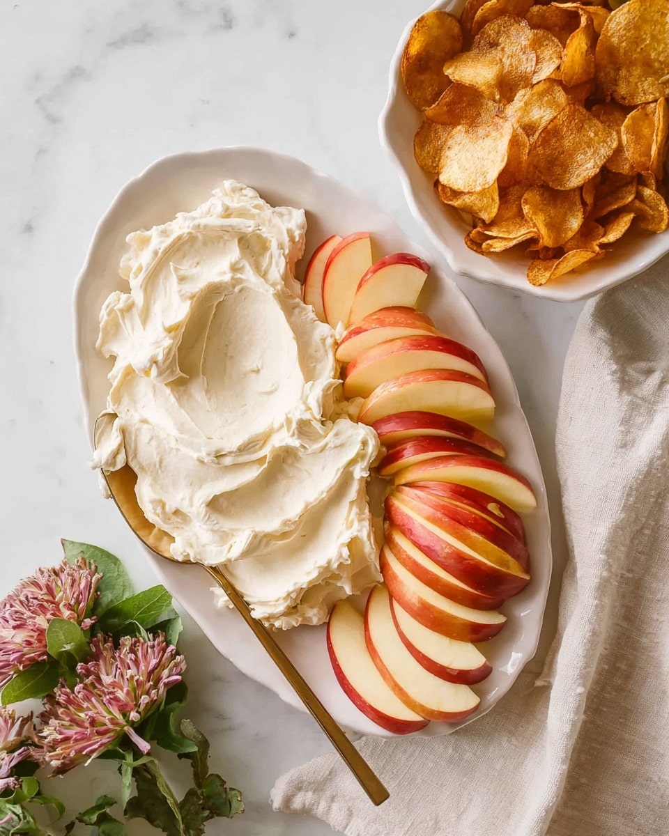 A white oval plate holds two main layers: on the left side, there is a thick, creamy white dip with a smooth and slightly whipped texture, and on the right side, there are neatly arranged apple slices with red and yellow skin stacked in two semi-overlapping rows. A gold-colored spreading knife rests on the dip. In the upper right corner, a white bowl is filled with golden-brown crispy chips, some chips scattered around the bowl. The plate and bowl sit on a white marbled surface with some small green and pink flowers and a cream-colored cloth nearby. photo taken with an iphone --ar 4:5 --v 7