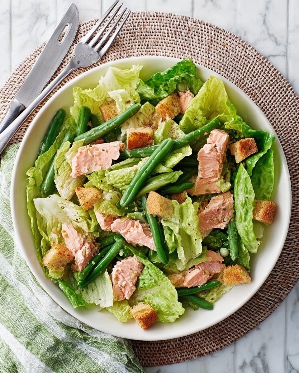 A fresh salad served in a white bowl, placed on a round woven mat over a white marbled surface. The salad has multiple layers: the bottom layer is large, bright green romaine lettuce leaves arranged around the bowl; scattered over the lettuce are pink pieces of cooked salmon; bright green whole beans lie on top, adding a long, slim texture; small golden-brown croutons are spread evenly around the salad, adding crunch. To the side of the bowl, a fork and knife rest on a striped green and white napkin. Photo taken with an iphone --ar 4:5 --v 7