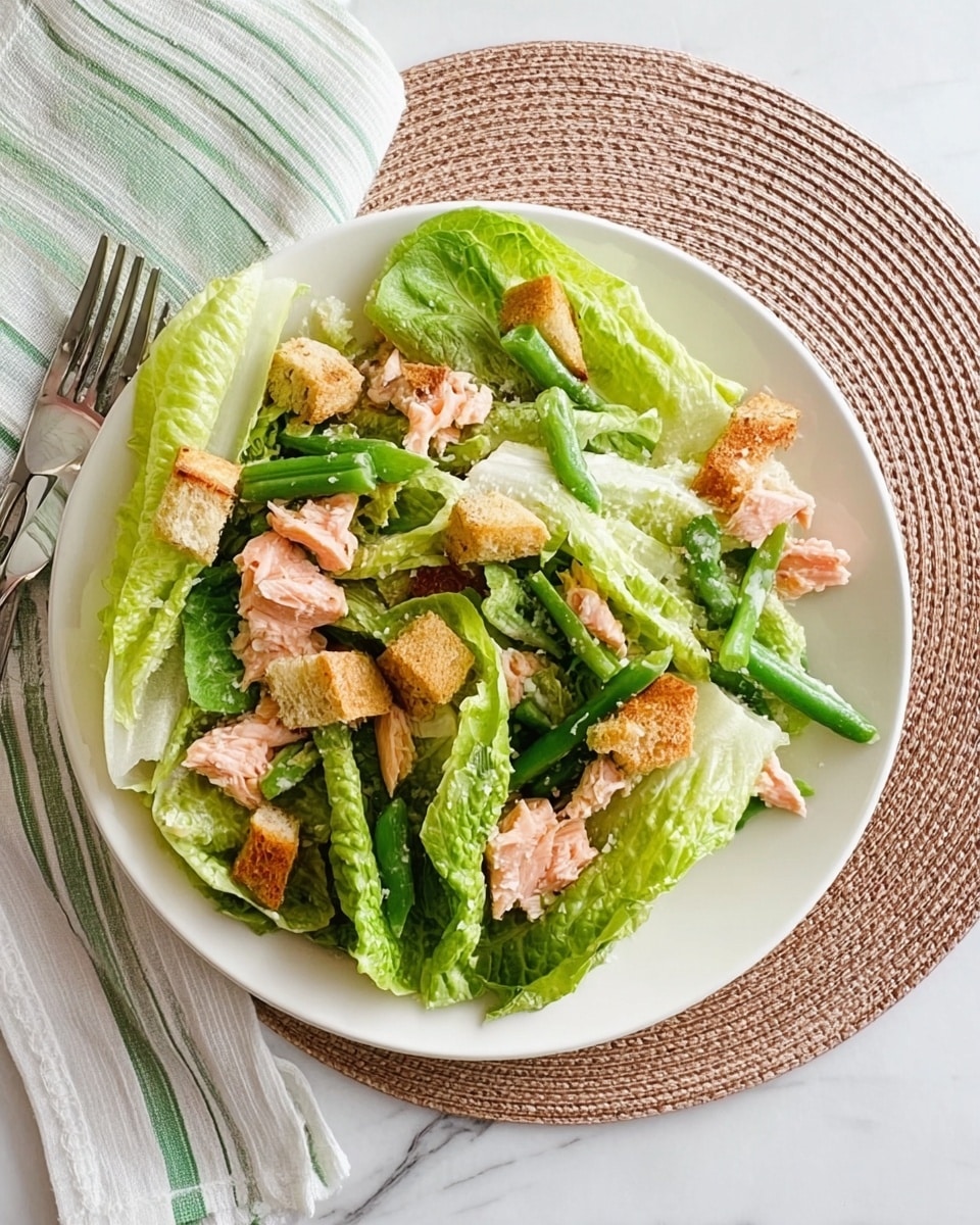 A white plate filled with a fresh salad placed on a white marbled surface with a round woven placemat underneath. The salad has several large, bright green romaine lettuce leaves layered unevenly around the plate, mixed with small pinkish pieces of cooked salmon. Thin, long green chili peppers are scattered on top and throughout the salad. Small light brown croutons are spread evenly over the lettuce and salmon, adding texture. To the left side of the plate, there is a folded green and white striped cloth napkin with a set of silver fork and knife resting on top. Photo taken with an iphone --ar 4:5 --v 7
