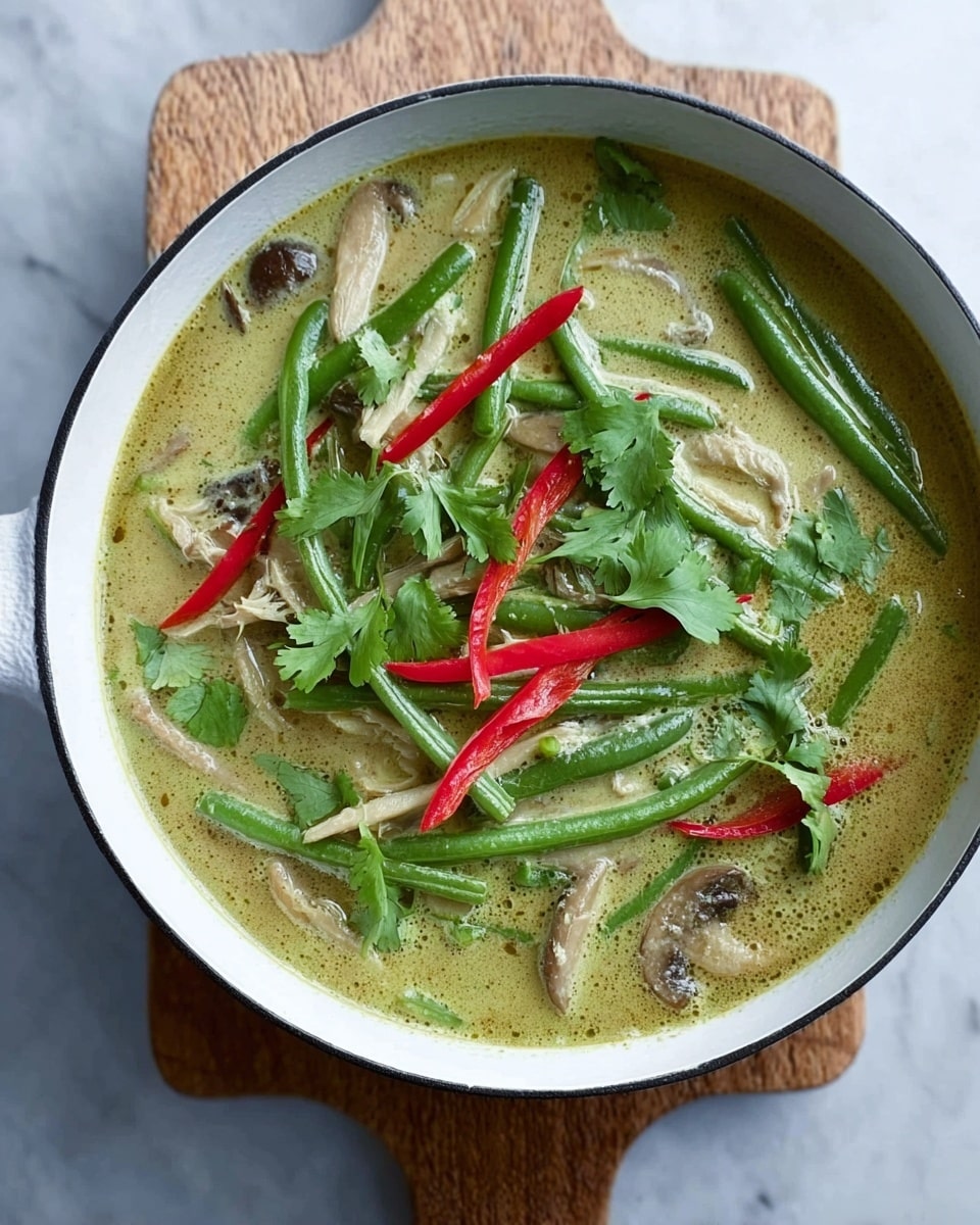 A white bowl filled with a creamy green curry soup that has a smooth, slightly oily surface. Inside the soup, there are bright green long beans scattered across, along with thin slices of light brown mushrooms, and thin red chili strips adding a pop of color on top. Fresh green cilantro leaves are spread unevenly, floating on the surface. The bowl rests on a white wooden board on a white marbled surface. Photo taken with an iphone --ar 4:5 --v 7
