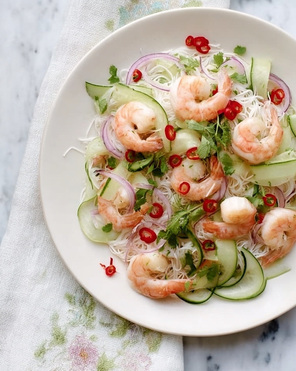 A white plate holds a fresh shrimp salad with three layers: the base is a mix of thin white rice noodles and thin purple-red onion slices, on top of which lie light pink cooked shrimp and pale green cucumber ribbons curled elegantly. Scattered throughout are bright red chili slices and small green herb leaves, adding pops of color and texture. The plate is placed on a white marbled surface partially covered by a light fabric with subtle floral patterns. photo taken with an iphone --ar 4:5 --v 7