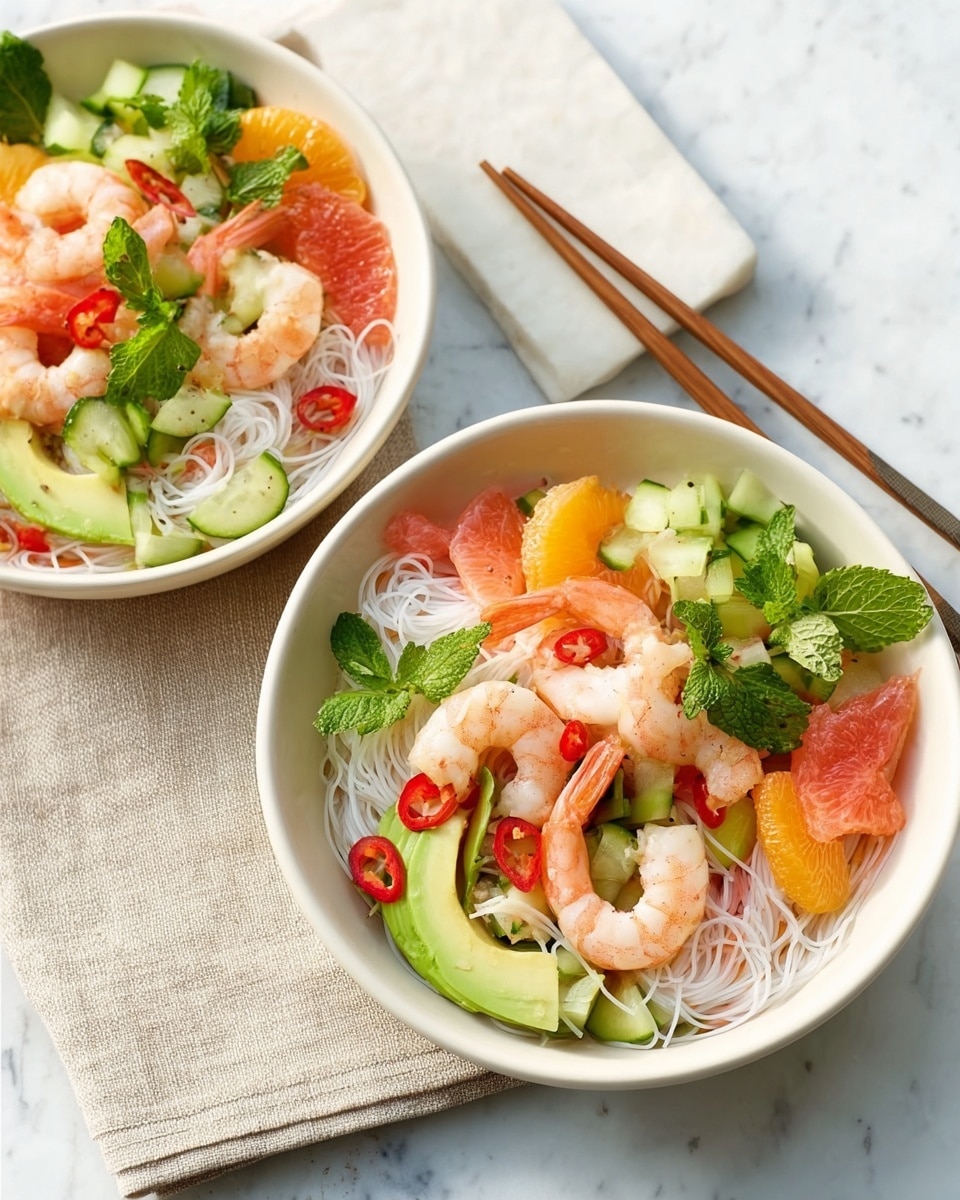 The image shows two white bowls filled with a colorful shrimp noodle salad placed on a white marbled surface. Each bowl has three main layers visible: the bottom layer consists of thin white rice noodles with a smooth texture, the middle layer has slices of light green avocado, thin orange carrot strips, and red chili slices adding brightness, and the top layer is decorated with plump pink shrimp, light yellow grapefruit segments, and green fresh herbs like cilantro and mint. The scene is completed with a folded beige cloth napkin and wooden chopsticks resting beside the bowls. Photo taken with an iphone --ar 4:5 --v 7