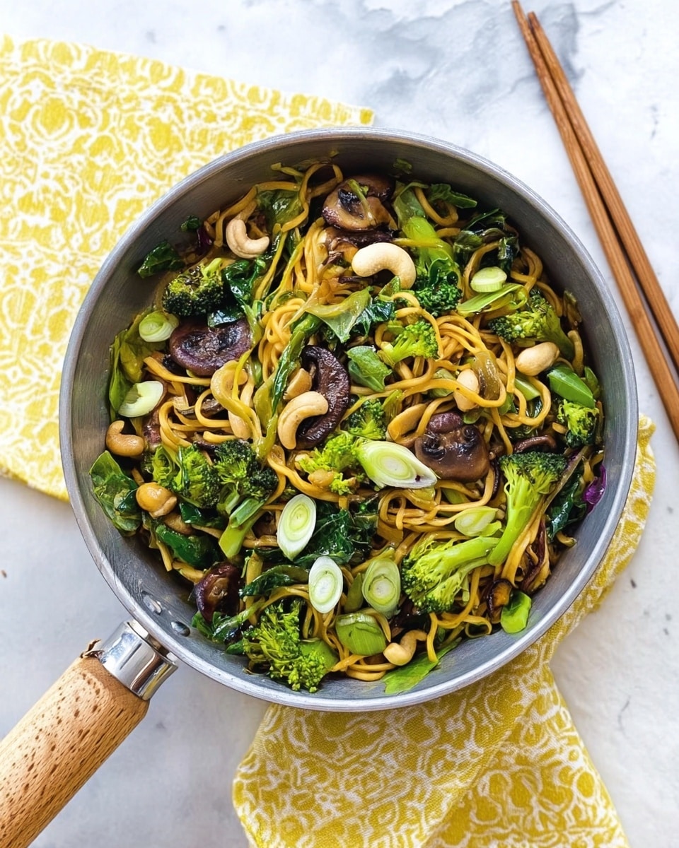 A metal pan containing a colorful noodle dish with two main layers visible: the bottom layer is yellow-brown noodles, and on top are green broccoli pieces, dark brown mushroom slices, and small white cashew nuts scattered evenly. There are also some spring onion rounds and leafy greens mixed in. The pan is placed on a white marbled surface with a folded yellow and white patterned cloth nearby, and a pair of wooden chopsticks rests to the side. photo taken with an iphone --ar 4:5 --v 7
