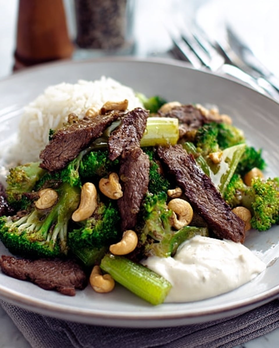 The dish features a white plate filled with a mixture of cooked food items. On the bottom layer, there are several dark brown strips of steak arranged in a slightly scattered way. Mixed throughout and on top of the steak are bright green pieces of broccoli and lighter green cooked celery sticks. Light brown toasted cashew nuts are spread over the dish, adding texture and contrast. At the back of the plate, there is a small mound of white rice, soft and fluffy in appearance. To the right side of the plate, a dollop of creamy white sauce creates a smooth shape. The plate sits on a white marbled surface, and a fork, knife, and pepper shaker are blurred in the background. Photo taken with an iphone --ar 4:5 --v 7