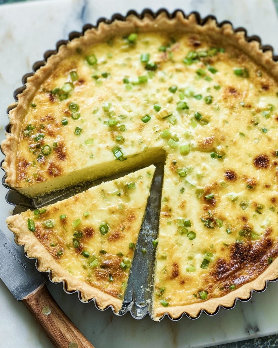 A golden-brown quiche with a crisp, slightly raised crust in a fluted tart pan, filled with a creamy yellow egg mixture that has small bright green pieces of chopped scallions scattered throughout; the top is lightly browned in spots. One slice is being lifted, showing the smooth texture of the inside. A knife with a wooden handle lies next to the quiche on a white marbled surface. Photo taken with an iphone --ar 4:5 --v 7