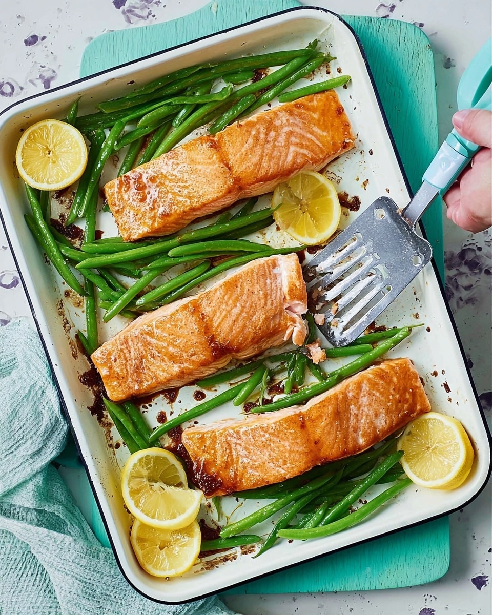 The image shows a white baking tray with three golden-brown cooked salmon fillets placed in the center, their texture crispy on top. Around the salmon are bright green steamed green beans arranged along the edges on both the left and right sides. There are also four lemon wedges with a yellow color placed in the bottom right corner and near the green beans on the top. A silver spatula with a blue handle rests on the right side of the tray, some of it touching the salmon. The tray sits on a white marbled surface, with a teal-colored cutting board beneath it, and the corner of a woman's hand holding a fork appears on the left side. Photo taken with an iphone --ar 4:5 --v 7