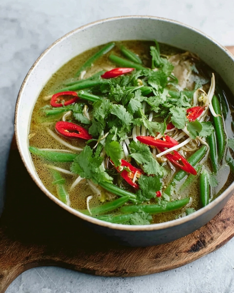 A white bowl filled with a layered soup, with a light green broth as the base, topped with whole green beans, sliced red chili peppers, fresh cilantro leaves scattered on top, and some thin, light-colored noodles or bean sprouts partly submerged in the broth. The bowl is placed on a rustic wooden board, set against a white marbled textured surface. photo taken with an iphone --ar 4:5 --v 7