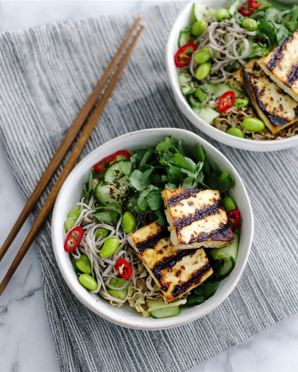 Two white bowls filled with fresh, colorful salad sit on a white marbled surface with a gray and white striped cloth beneath them. Each bowl contains two slices of grilled tofu with dark grill lines, placed on top of a bed of mixed greens, light green cucumber slices, bright green edamame, thin light gray noodles, and red chili pieces. Fresh green herbs add a pop of color on top. A pair of wooden chopsticks rests next to the bowls, aligned on the striped cloth. In the background, a woman's hand delicately holds one of the bowls. Photo taken with an iphone --ar 4:5 --v 7