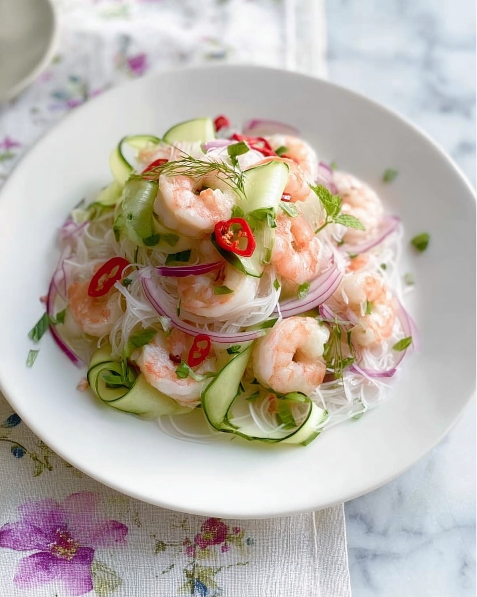 A white plate holds a shrimp salad with several layers: the bottom layer consists of thin white rice noodles, followed by pink shrimp scattered on top. Around the shrimp are pale green cucumber ribbons curled into loops, with thin slices of light purple-red onion spread evenly across the dish. Bright red chili slices and small fresh green cilantro leaves are sprinkled throughout, adding color and texture. The plate sits on a white marbled surface partially covered with a light cloth that has a delicate floral pattern. Photo taken with an iphone --ar 4:5 --v 7