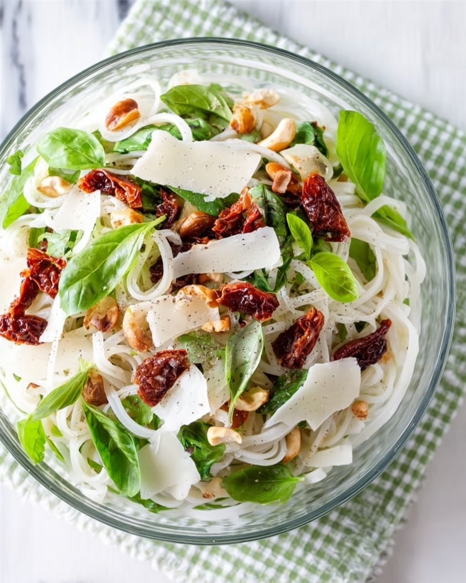 The image shows a clear glass bowl filled with a layered pasta salad placed on a white marbled surface with a green and white checkered cloth underneath. The bottom layer consists of white rice noodles, loose and slightly curled. On top of this is a mix of bright green basil leaves and scattered halves of dark, dry sun-dried tomatoes. Thin, broad shavings of off-white cheese are spread across the salad, along with small pieces of chopped light-colored nuts. The dish is finished with a light sprinkle of black pepper, adding texture and contrast to the fresh ingredients. Photo taken with an iphone --ar 4:5 --v 7