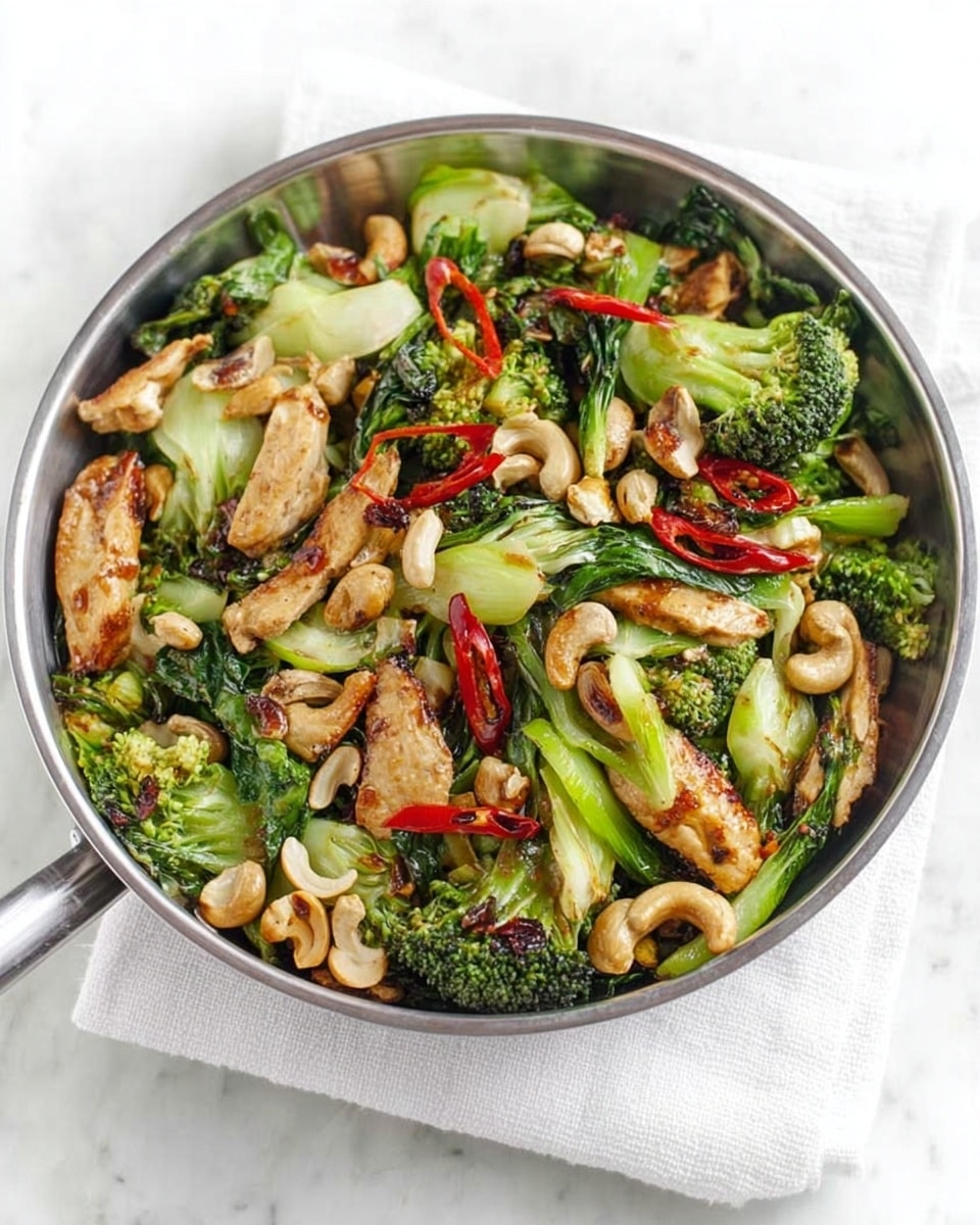 A large round metal bowl filled with a colorful stir-fry of green broccoli florets, light green bok choy pieces with darker green tops, golden brown grilled chicken strips, white cashew nuts scattered throughout, and thin slices of red chili peppers on top for color. The bowl is placed on a white folded towel, resting on a white marbled surface. photo taken with an iphone --ar 4:5 --v 7