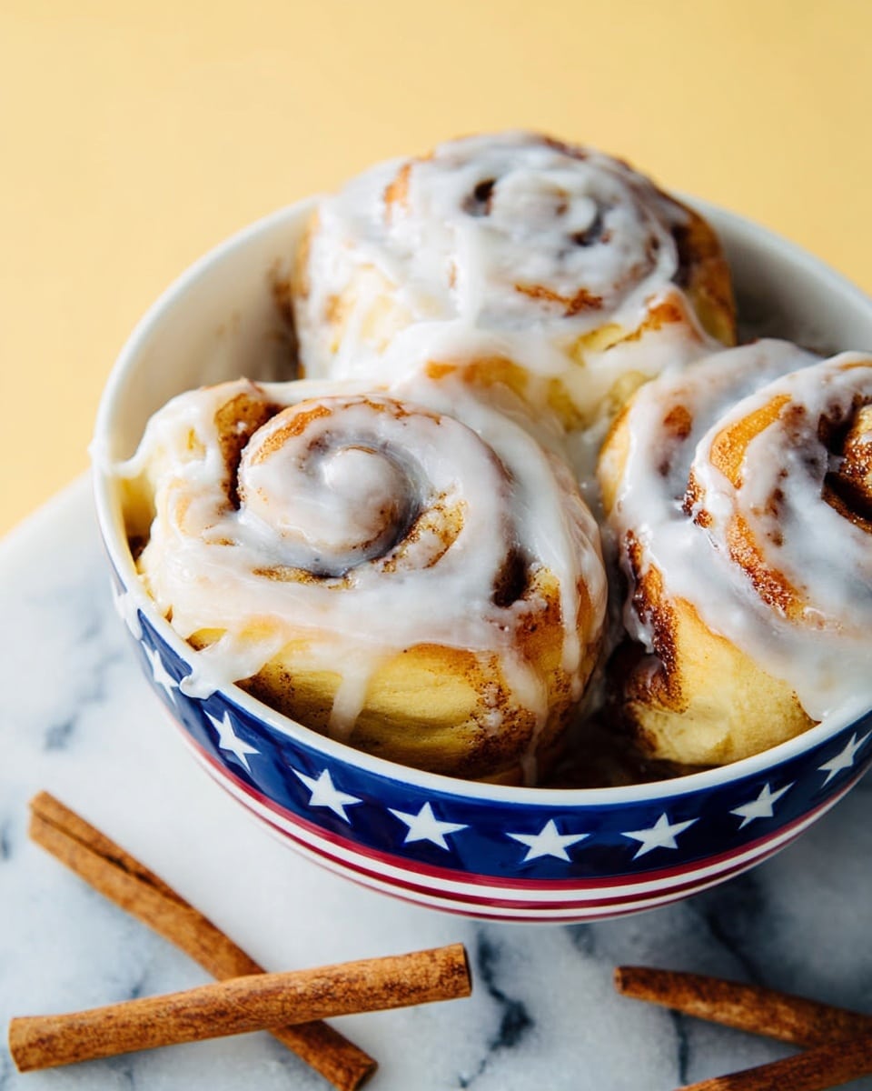 A close-up view of five cinnamon rolls with thick white icing spread unevenly over the top, showing golden-brown baked dough with visible cinnamon swirls inside each roll; the rolls are arranged in a round white bowl decorated with blue stars and stripes, placed on a white marbled texture surface with two cinnamon sticks nearby; the rolls look soft and freshly baked with a slightly shiny glaze from the icing photo taken with an iphone --ar 4:5 --v 7