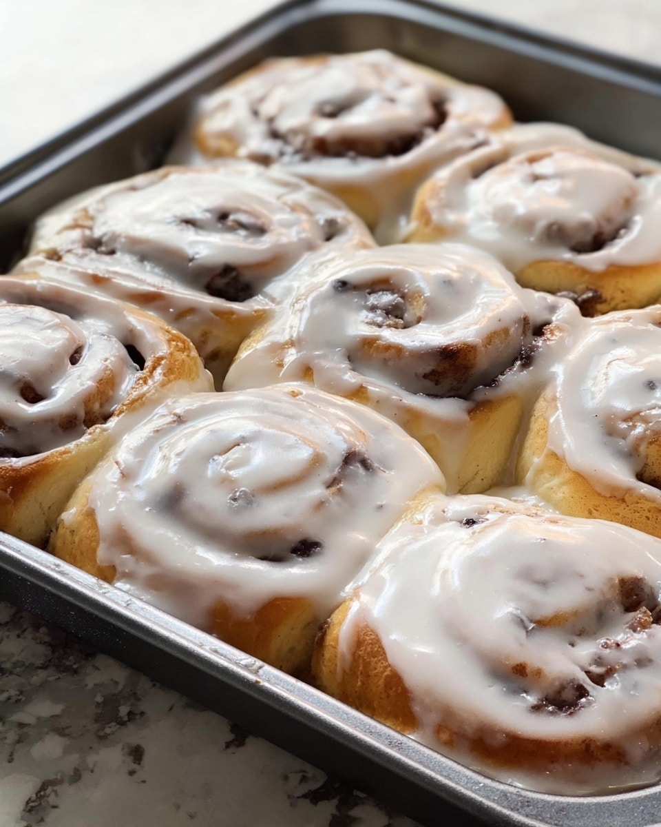 A metal baking pan filled with nine cinnamon rolls that are thick and soft, each roll has a light golden brown base with visible swirls of cinnamon inside, topped generously with a smooth, white glaze that covers the rolls almost like a creamy blanket, the glaze is shiny and slightly runny, dripping slightly down the sides of the rolls, the pan sits on a white marbled texture surface. photo taken with an iphone --ar 4:5 --v 7