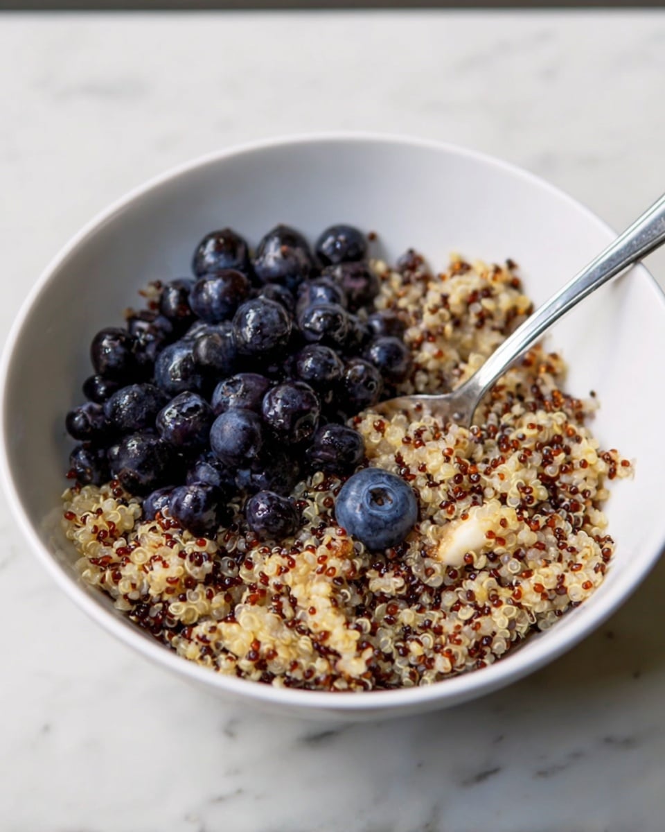 A white bowl filled with a base layer of cooked quinoa, which is a mix of white, red, and black grains giving a speckled and soft texture. On top of the quinoa, there is a small heap of fresh blueberries that are dark blue and shiny, contrasting with the light color of the quinoa. A silver spoon is placed inside the bowl resting on the right side, partially submerged in the quinoa and blueberries. The bowl sits on a white marbled surface. photo taken with an iphone --ar 4:5 --v 7