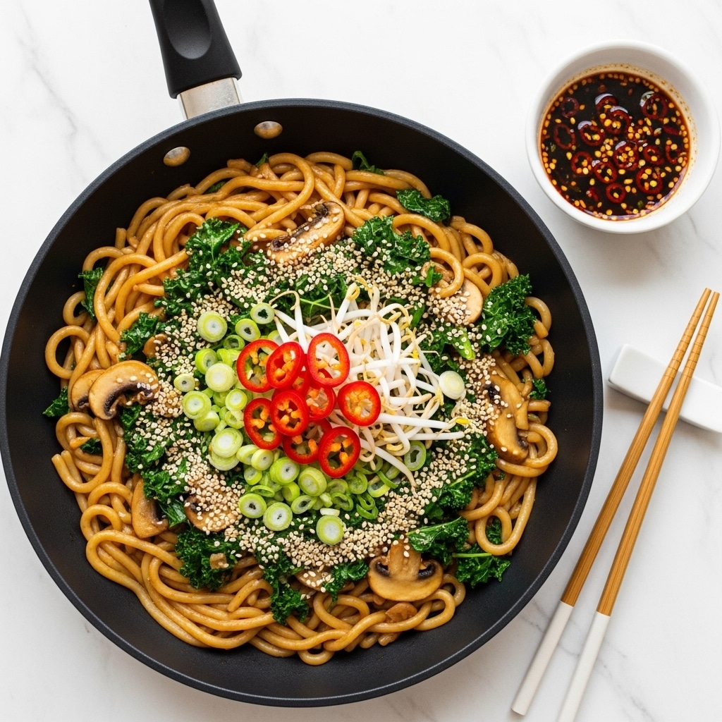 In a black pan, thick udon noodles form the main layer, cooked light brown and mixed with wilted dark green kale pieces and sliced light tan mushrooms; scattered thin slices of bright red chili peppers and chopped fresh green onions add spots of color, topped with light beige sesame seeds and a few white bean sprouts for texture. Next to the pan on a white marbled surface, there is a small white bowl filled with dark soy sauce sprinkled with red chili flakes. A pair of light wooden chopsticks rests on a white chopstick holder beside the bowl. The photo taken with an iphone --ar 4:5 --v 7