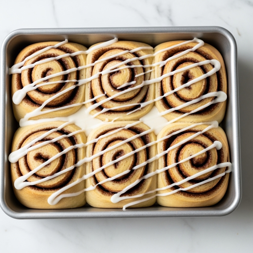 The image shows a close-up view of six cinnamon rolls in a silver baking tray. Each roll has multiple spiral layers: a soft, light golden dough forming the base layer, and a dark brown cinnamon filling layer creating the swirl pattern inside. The tops of the rolls are generously drizzled with a smooth, white icing that shines slightly and drips down the sides. The tray is placed on a white marbled surface, highlighting the warm colors of the cinnamon rolls. photo taken with an iphone --ar 4:5 --v 7
