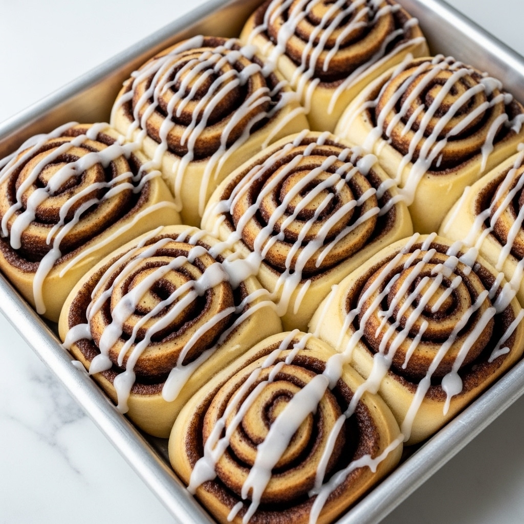 The image shows a close-up of six cinnamon rolls in a silver baking tray, each roll with a spiral shape made of light golden brown dough and darker brown cinnamon filling. The rolls are topped with a smooth white icing drizzled in wavy lines across the top, giving a glossy, creamy texture. The tray sits on a white marbled surface, with visible soft and fluffy texture on the rolls’ edges where dough layers overlap. The icing highlights the swirls while also pooling slightly between some rolls. photo taken with an iphone --ar 4:5 --v 7