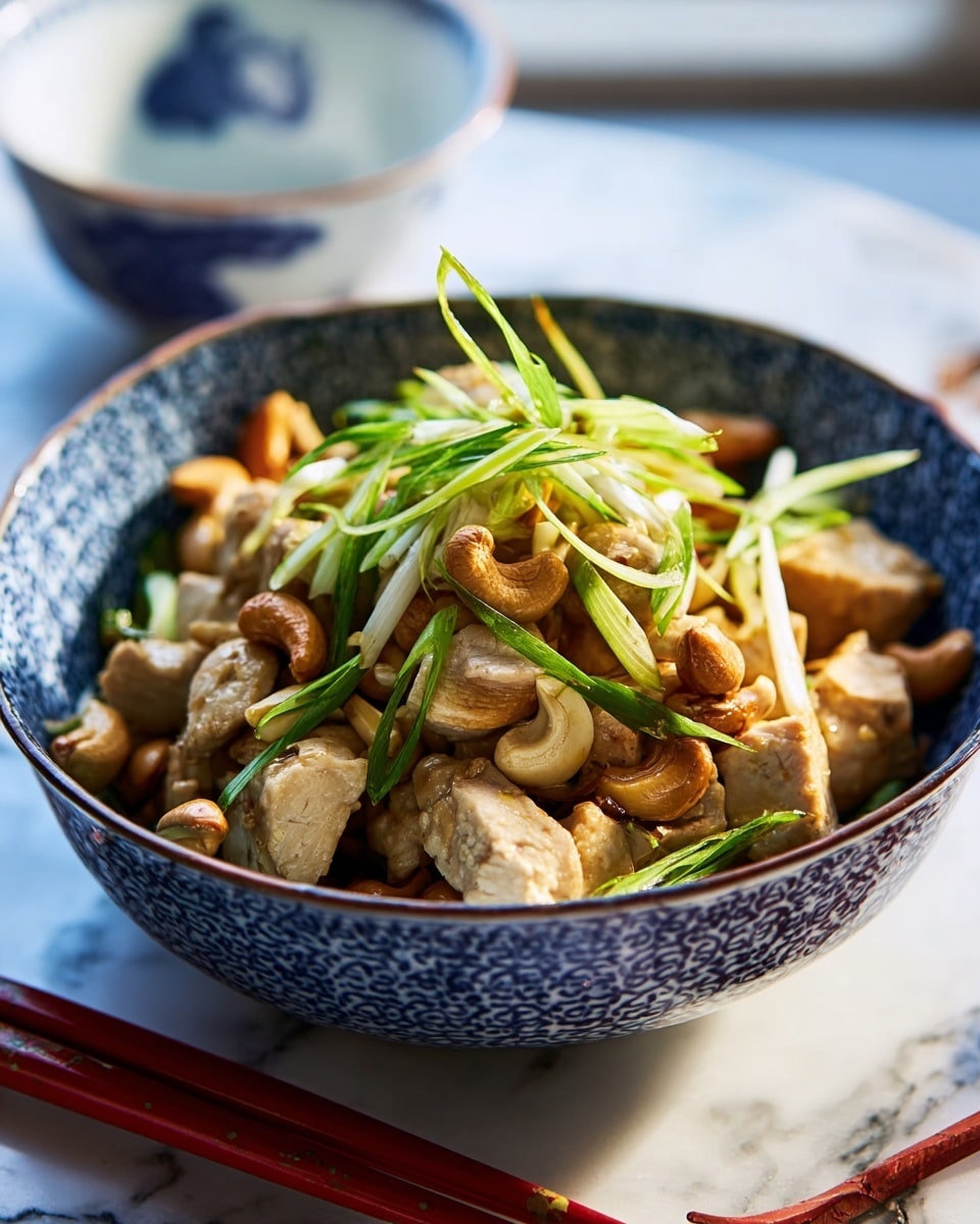 The image shows a blue and white patterned bowl filled with a dish made of bite-sized pieces of light brown cooked chicken, scattered with light golden cashew nuts, and topped with thin, long strips of bright green scallions placed in the center. The bowl sits on a white marbled surface with a red cloth partially visible beneath it, and wooden chopsticks rest beside the bowl. Photo taken with an iphone --ar 4:5 --v 7