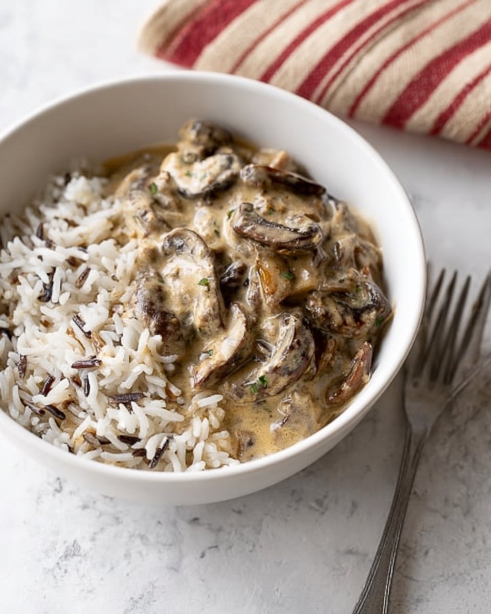 A white bowl filled with two layers, the bottom layer is white and brown grains of wild rice, and the top layer is creamy mushroom sauce with sliced brown mushrooms coated in a light beige sauce. The bowl is placed on a white marbled surface, next to a metal fork on the right side. Photo taken with an iphone --ar 4:5 --v 7