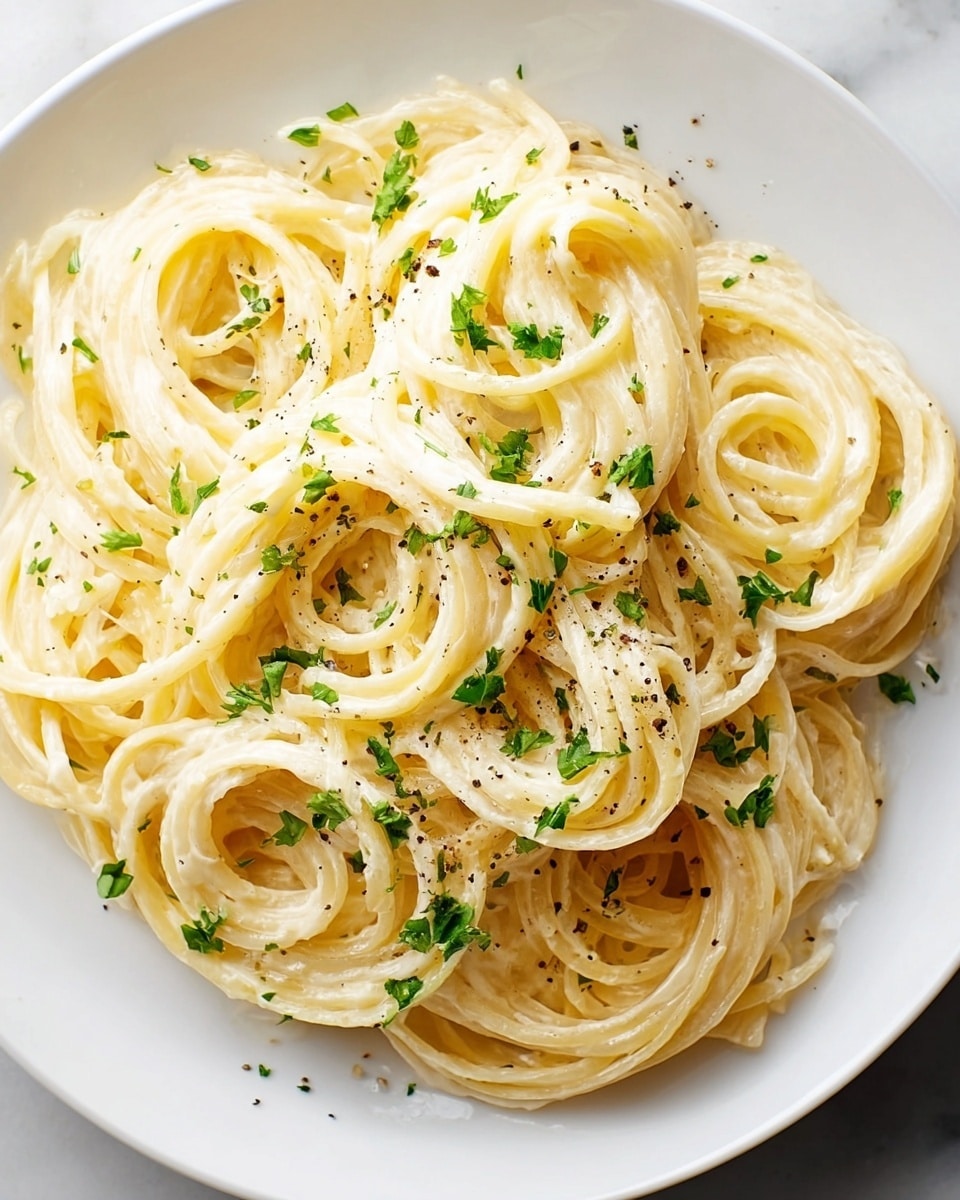 A close-up view of a white round plate filled with creamy spaghetti. The pasta is twirled into small nests piled closely together, coated evenly with a smooth, pale yellow sauce. There are small pieces of green herbs sprinkled over the top, adding a fresh contrast. Tiny specks of black pepper are scattered across the noodles, giving a textured look. The plate sits on a white marbled surface, enhancing the clean and simple presentation. photo taken with an iphone --ar 4:5 --v 7