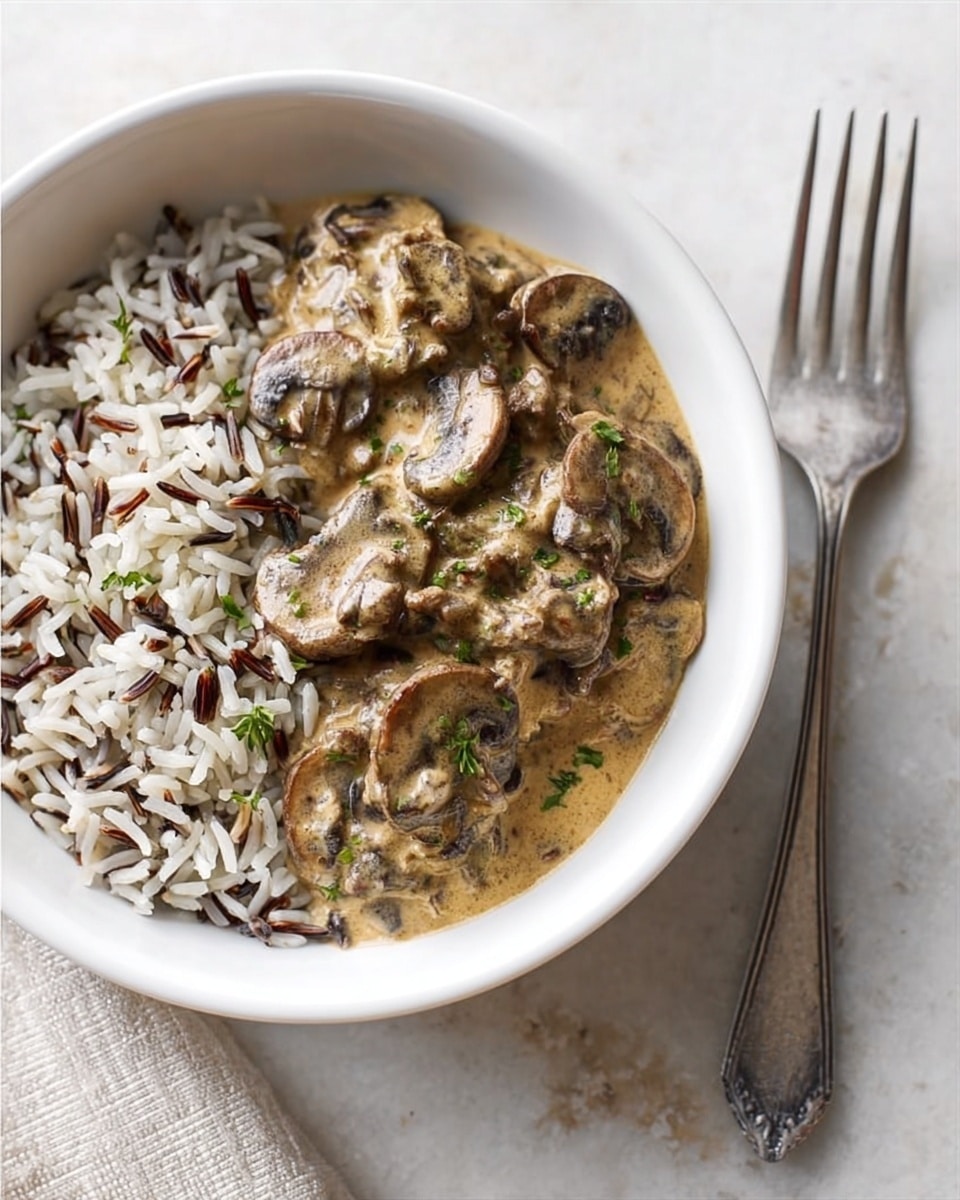 A white bowl with two main layers: the bottom layer is white and brown wild rice with a fluffy texture, filling half of the bowl; the top layer is a creamy, light brown mushroom sauce with visible slices of dark brown mushrooms mixed in, covering the rice and flowing slightly over the edges. The bowl is placed on a white marbled surface with a metal fork to the right side and a cloth with red and beige stripes in the background. Photo taken with an iphone --ar 4:5 --v 7
