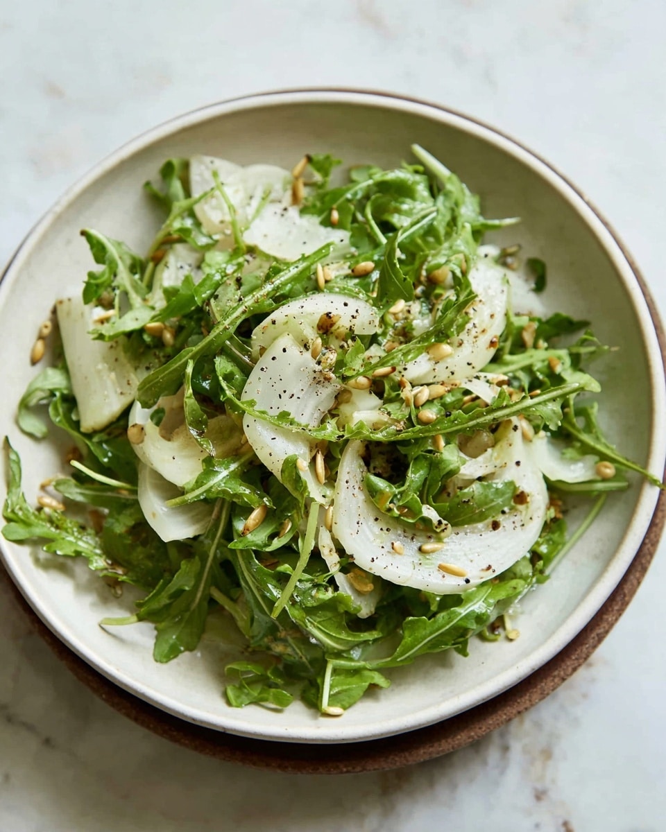 A simple green salad is shown in a shallow white bowl, filled with mixed green leaves like arugula and thinly sliced white fennel pieces spread throughout. The salad is topped lightly with a sprinkle of small, light brown seeds and cracked black pepper scattered unevenly, adding texture and contrast. The greens look fresh and slightly shiny as if lightly dressed with oil, and the bowl sits on a white marbled surface. Photo taken with an iphone --ar 4:5 --v 7