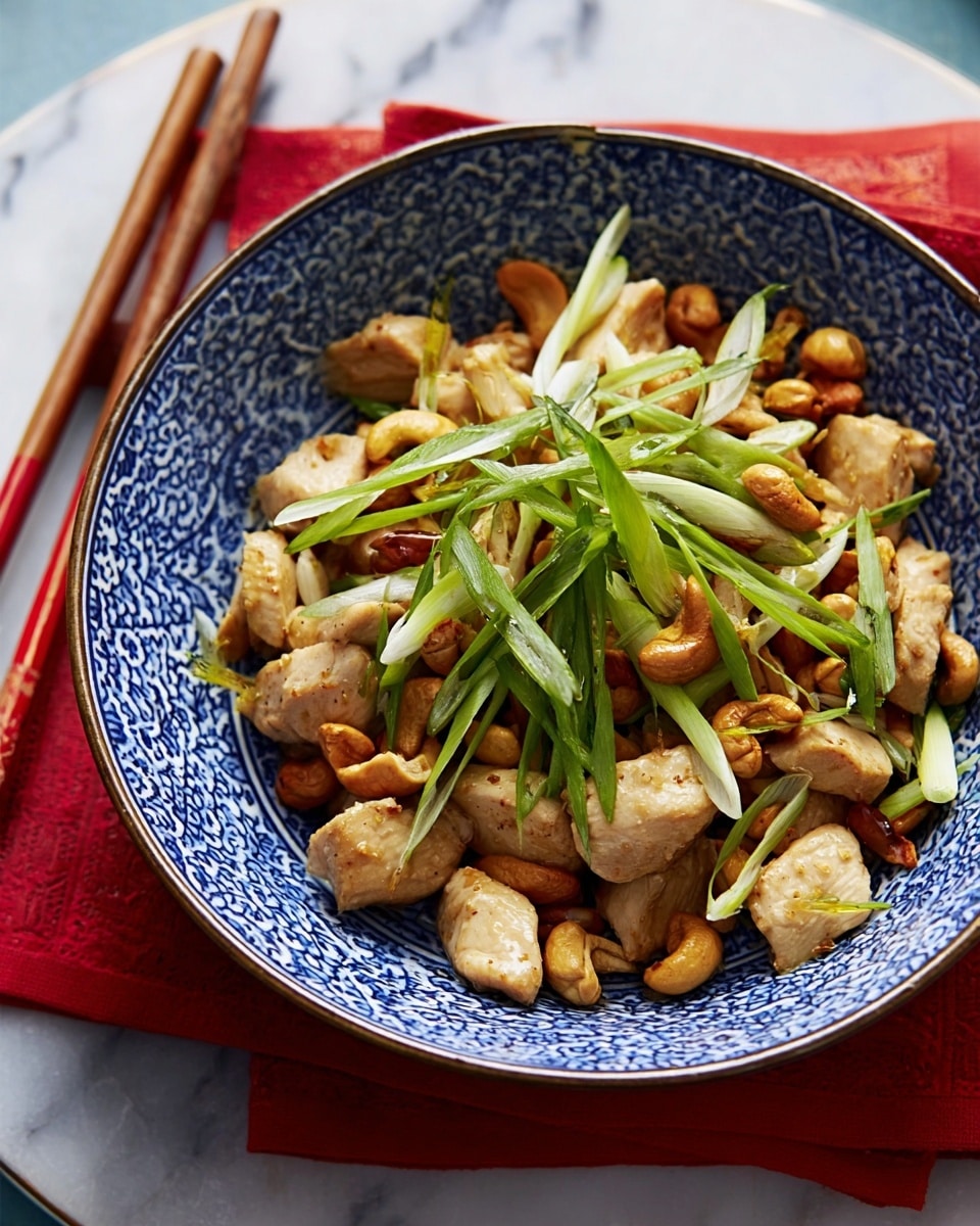 A close-up of a blue and white patterned bowl filled with a dish that has several layers: chunks of light brown cooked chicken form the base layer, topped with golden cashew nuts scattered around, and finished with thin, bright green strips of scallions placed on top. The bowl sits on a white marbled surface with chopsticks resting next to it and a small white bowl with blue patterns in the background. The lighting is natural, highlighting the textures of the chicken and nuts, photo taken with an iphone --ar 4:5 --v 7