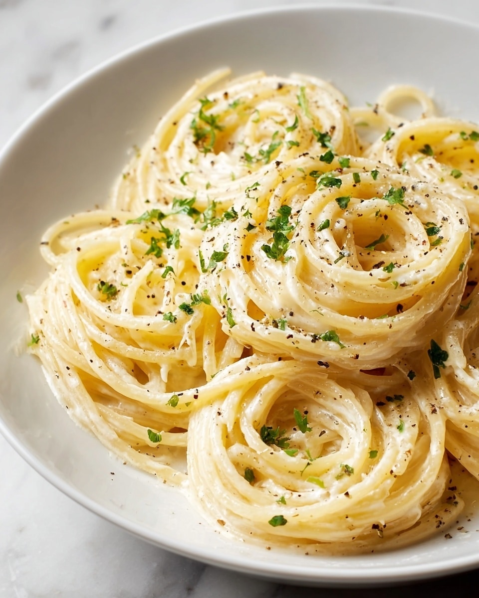 A white plate filled with creamy spaghetti arranged in loose, round swirls, showing thick, smooth noodles covered evenly with a glossy, pale yellow cream sauce. Small bits of fresh green parsley are sprinkled over the pasta, with tiny specks of black pepper adding contrast. The plate sits on a white marbled surface, giving a clean and bright look. photo taken with an iphone --ar 4:5 --v 7