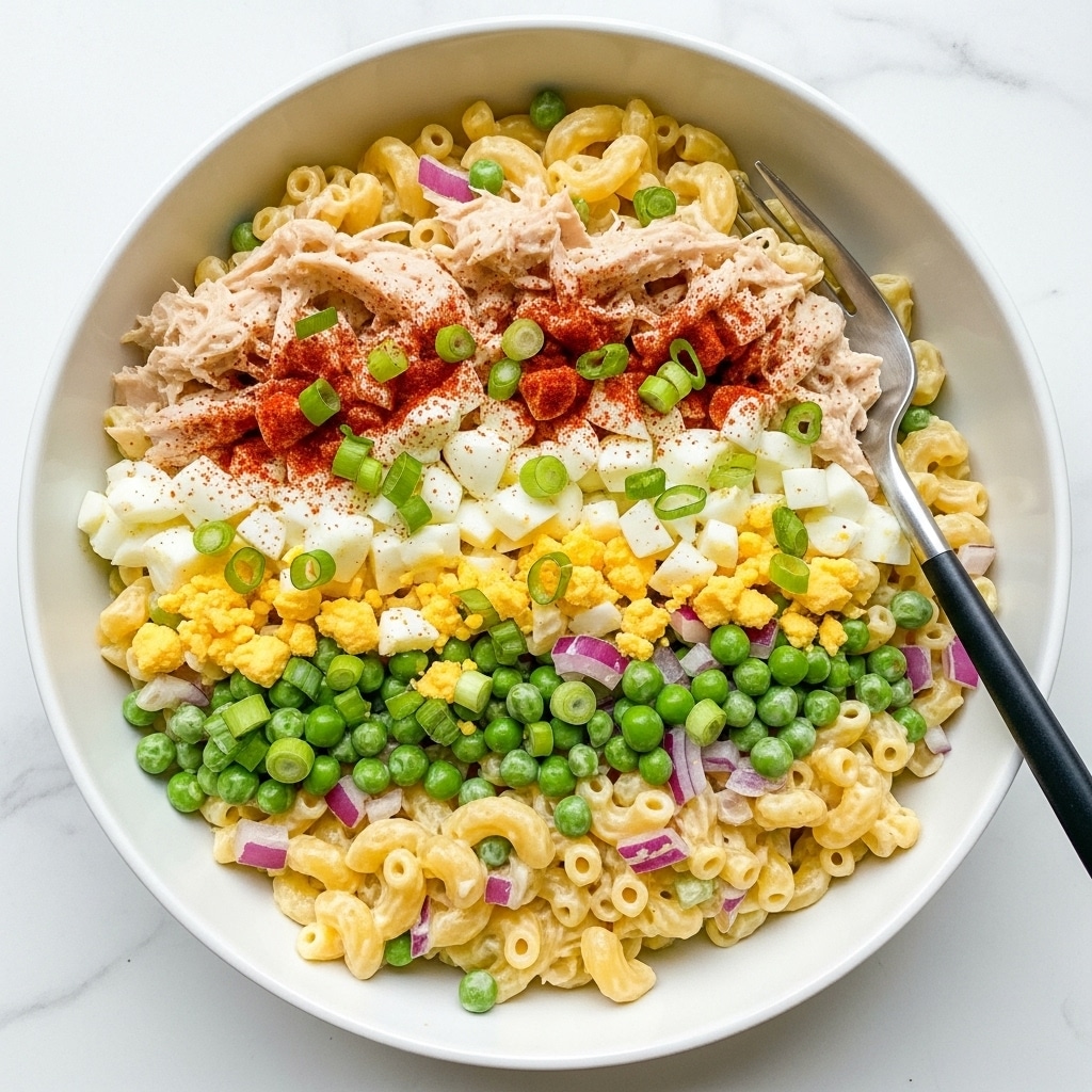 A close-up view of a white bowl filled with creamy macaroni salad, showing about three layers of small elbow macaroni coated in a pale yellow dressing. Mixed within are chunks of white boiled egg with a soft texture, green peas, small pieces of celery, and bits of red onion adding purple and green touches. Small chunks of white chicken are scattered throughout, and the salad is lightly sprinkled with reddish paprika and chopped green onions on top. A silver fork with a black handle lies inside the bowl on the right side, all sitting on a white marbled surface. photo taken with an iphone --ar 4:5 --v 7