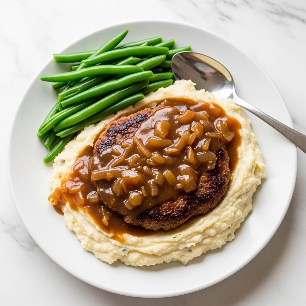 A white plate on a white marbled surface holds a meal with three main parts. At the base there is a smooth, creamy layer of mashed potatoes, white in color with light texture. On the right side of the plate, two pieces of browned meat covered with a glossy, thick brown gravy with visible pieces of cooked onions sit on top of the mashed potatoes. On the left side, there is a small pile of bright green, cooked green beans arranged neatly. A silver spoon rests on the right edge of the plate. photo taken with an iphone --ar 4:5 --v 7
