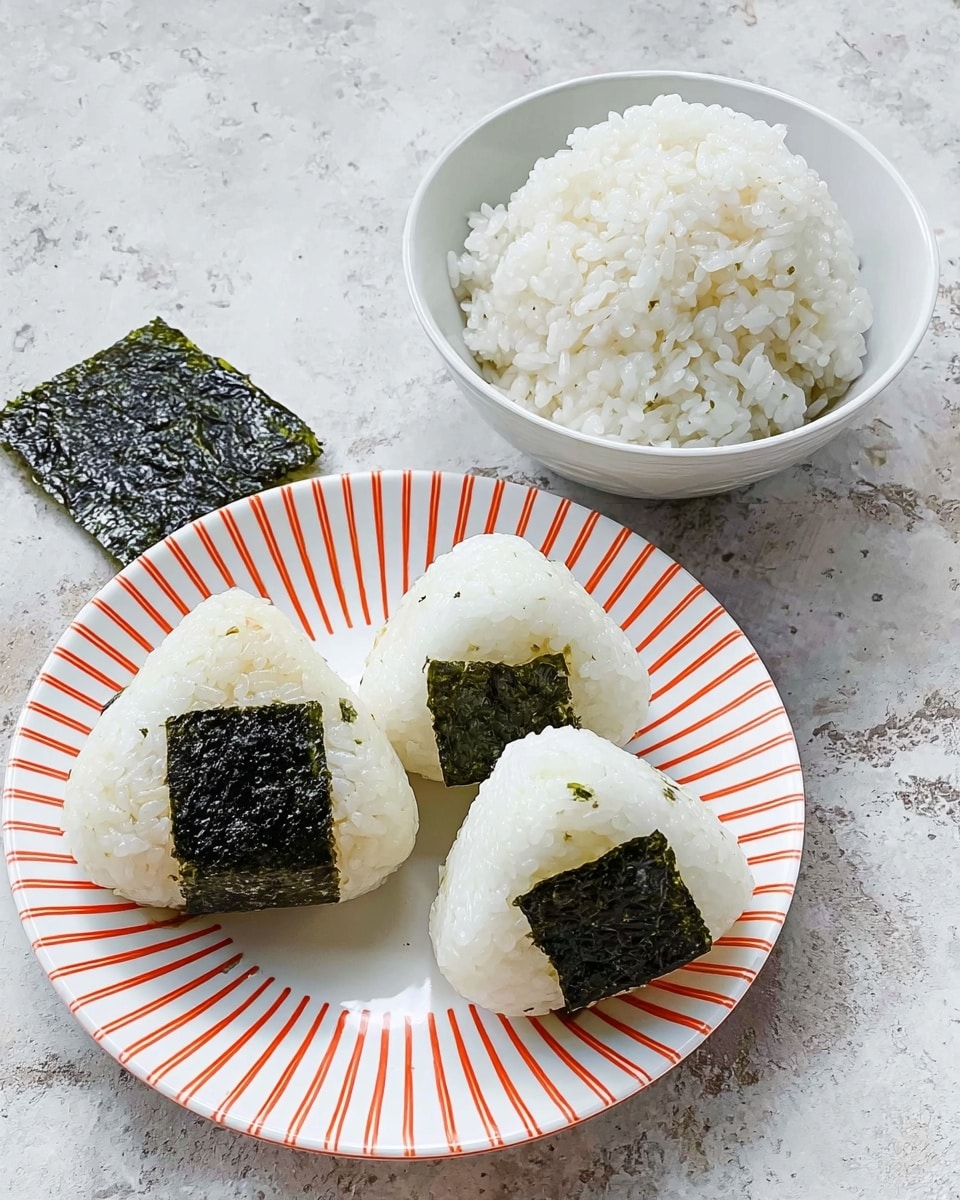 The image shows three triangular rice balls with a small dark green square of seaweed placed in the center on top, arranged on a white plate with thin orange diagonal stripes. Next to the plate, there is a white bowl filled with plain white rice, and a single piece of dark green seaweed sits on the white marbled surface beside the bowl. The background is a white marbled texture. photo taken with an iphone --ar 4:5 --v 7