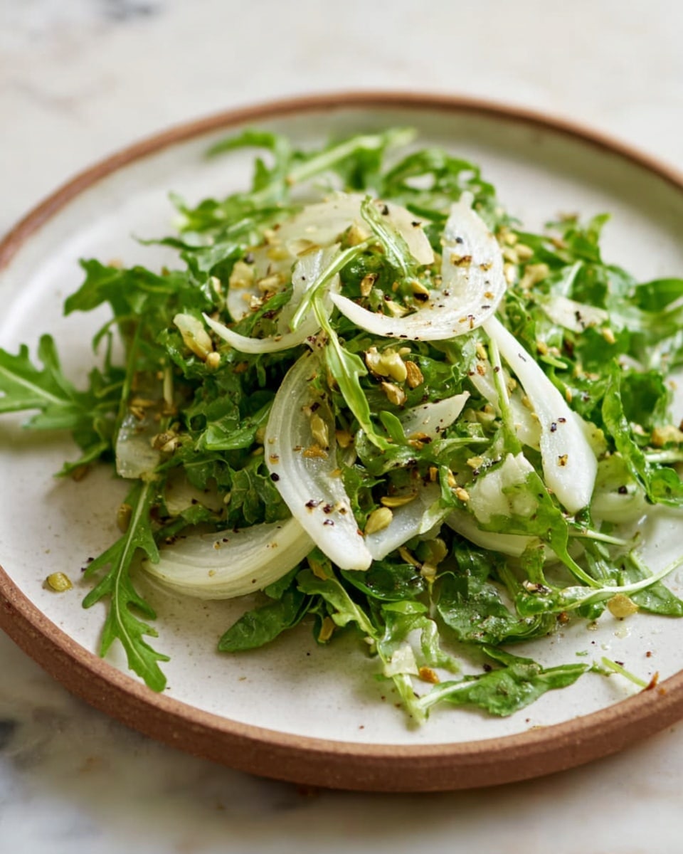 A fresh salad is placed on a round white plate, showing two main layers: a base of bright green leafy arugula with a slightly rough texture, topped with thin, pale white slices of fennel that look soft and layered. Scattered on top are small, light brown seeds, adding a crunchy texture, and the entire salad is lightly sprinkled with black pepper. The plate sits on a white marbled textured surface. Photo taken with an iphone --ar 4:5 --v 7