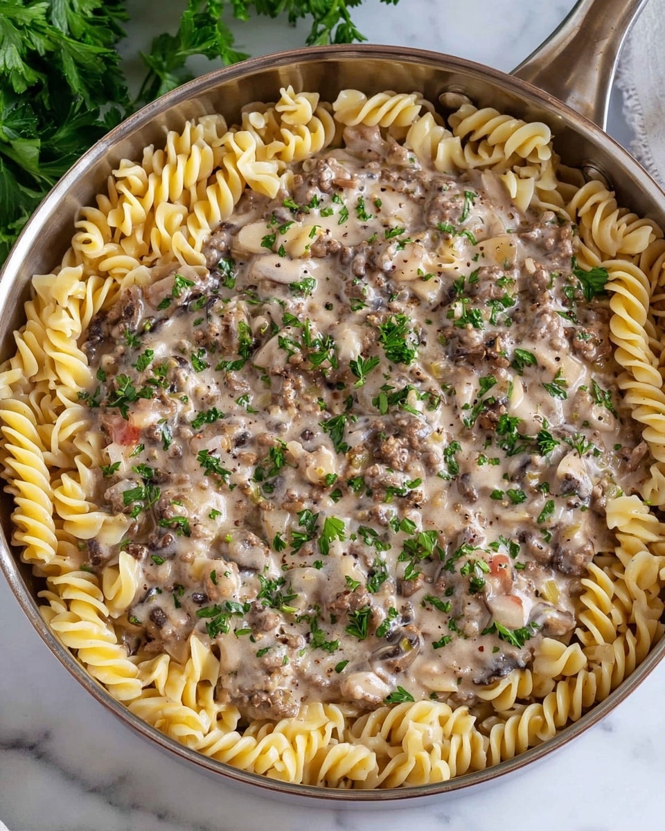 A top-down view of a white pan filled with two layers: the bottom layer is light beige spiral pasta arranged around the edges of the pan, creating a ring shape, while the center is filled with a creamy sauce with ground beef, diced onions, and bits of mushrooms, topped with green chopped herbs scattered evenly on the meat and sauce. The pan handle extends to the right side, and the pan sits on a white marbled surface with hints of green leaves in the background. photo taken with an iphone --ar 4:5 --v 7