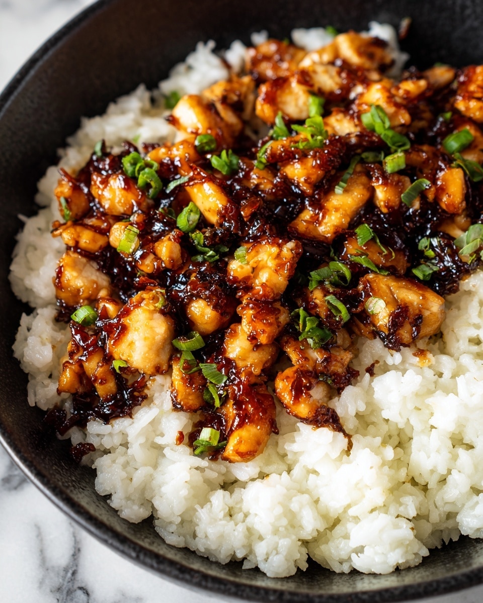 The dish shows a black bowl filled with two layers: the bottom layer is soft, white rice with a slightly sticky texture, while the top layer has glossy, caramelized chunks of chicken with a dark brown sauce drizzled over them. Small green pieces of chopped herbs and scallions are scattered on the chicken, adding bright green contrast. The background has a white marbled texture, making the colors of the food stand out clearly. Photo taken with an iphone --ar 4:5 --v 7