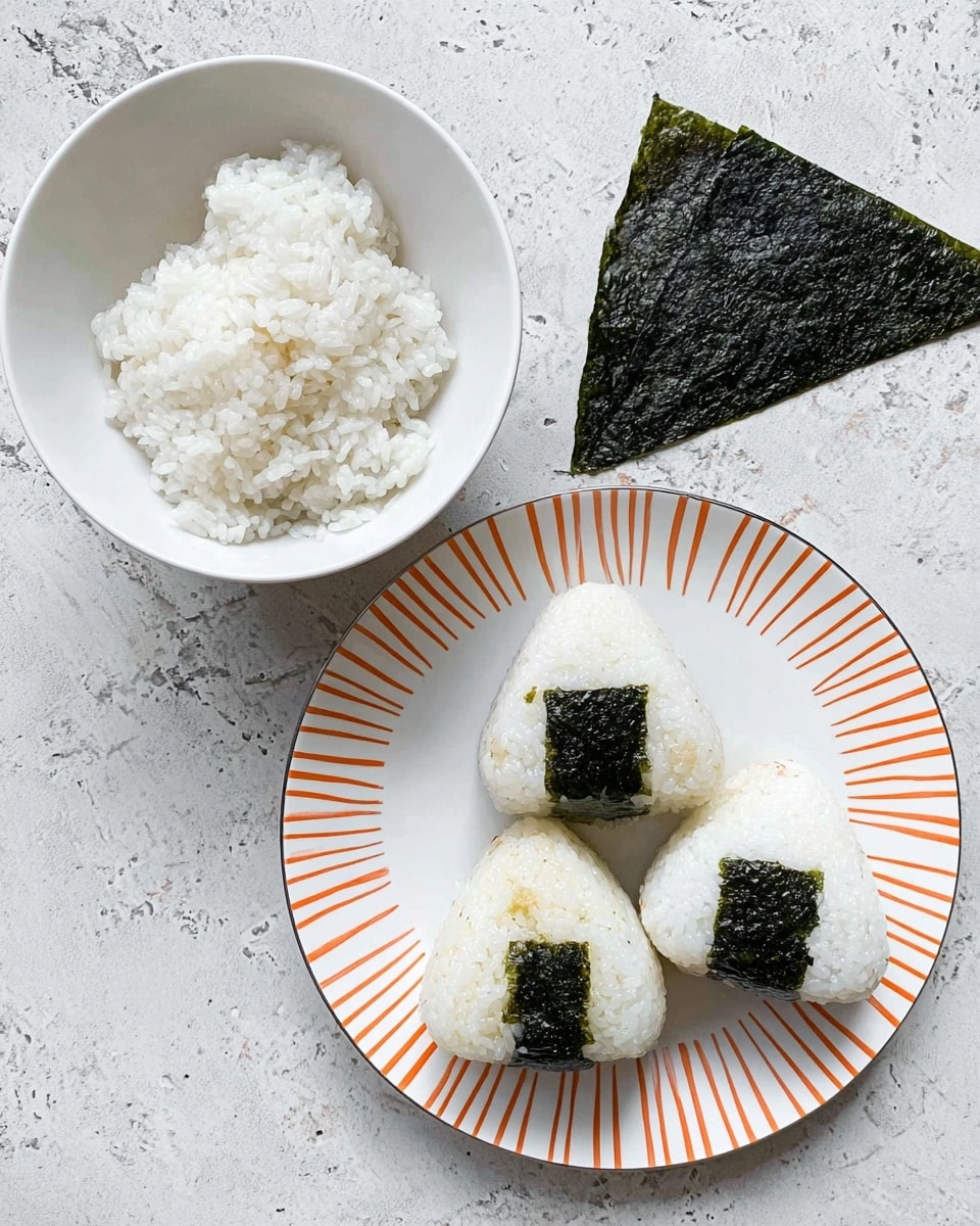 A white plate with orange lines holds three triangular rice balls, each topped with a small square of dark green seaweed on their upper part, giving a contrast to the white rice. To the left, there is a white bowl filled with plain white cooked rice, and above it lies a single rectangular piece of dark green seaweed. All items rest on a white marbled textured surface. photo taken with an iphone --ar 4:5 --v 7