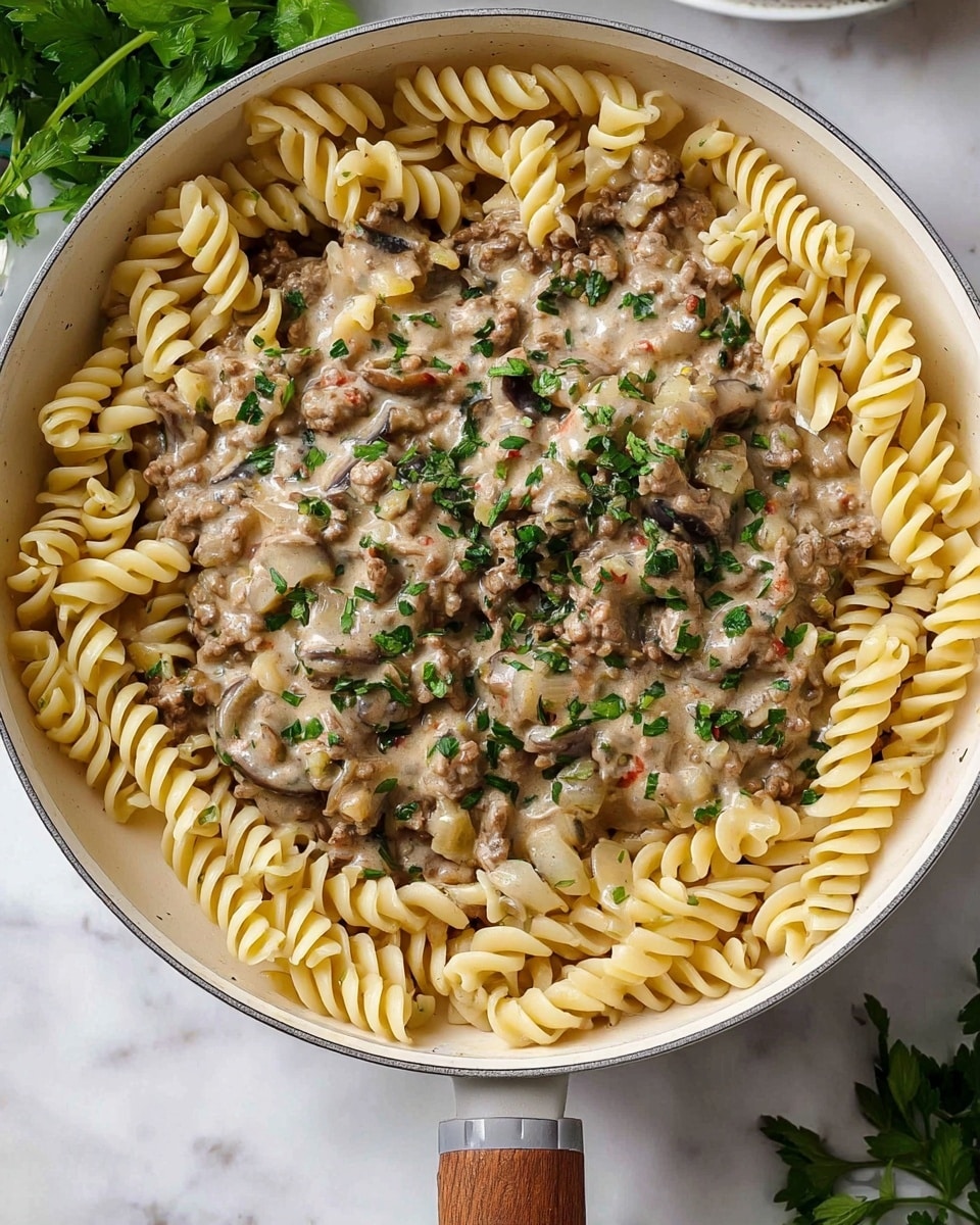 A round pan filled with two main layers; the bottom layer is a bed of light yellow twisted pasta spirals arranged in a circle along the edge of the pan, leaving the center empty. On top, there is a creamy mixture of ground beef, small chopped mushrooms, translucent onions, and bits of tomato, all coated in a pale grayish sauce with visible herbs and black pepper. The dish is sprinkled with finely chopped fresh green parsley, adding color contrast. The pan handle is visible on the top right, and the scene is set on a white marbled surface with some green leaves blurred in the background. Photo taken with an iphone --ar 4:5 --v 7